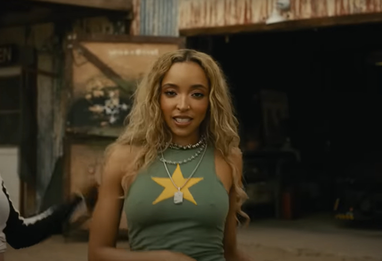 Tinashe is in an outdoor setting, wearing a sleeveless top with a large star graphic and layered necklaces. She is looking directly at the camera