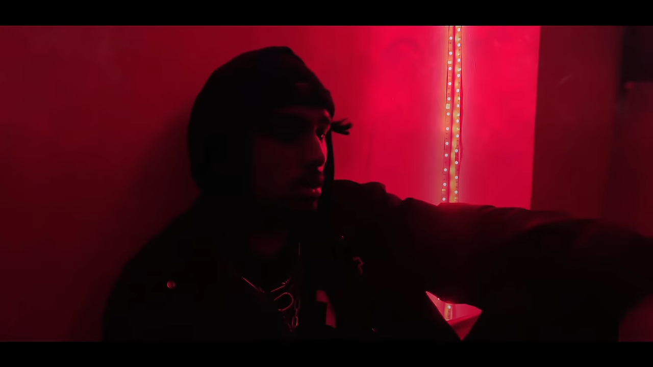 A person wearing a dark hoodie sits against a wall illuminated by red lighting. The atmosphere is intense and moody