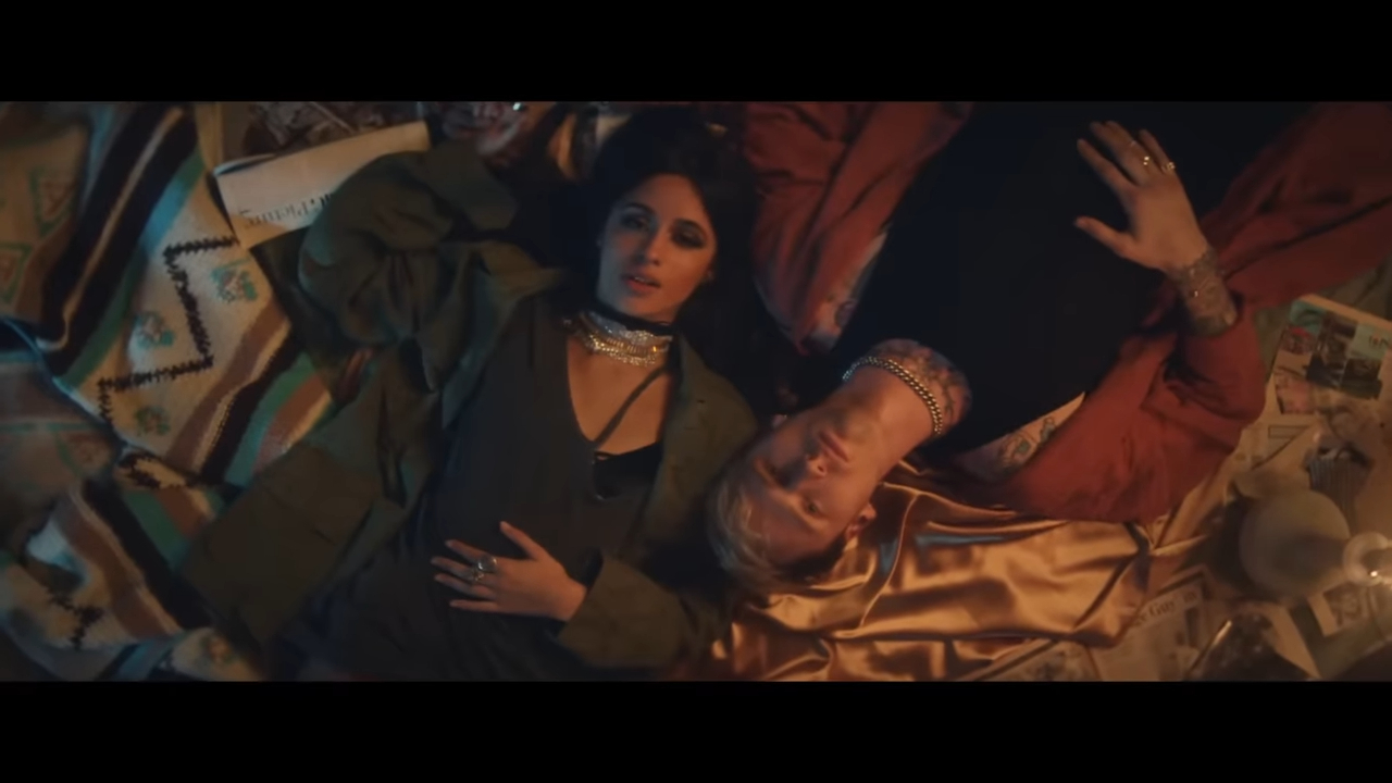 Camila Cabello and another person lie on a blanket, gazing upwards, surrounded by scattered papers and a candle, evoking a relaxed, intimate atmosphere