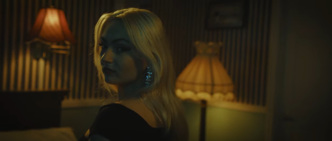 Woman with blonde hair and earrings in a dimly lit room glances over her shoulder