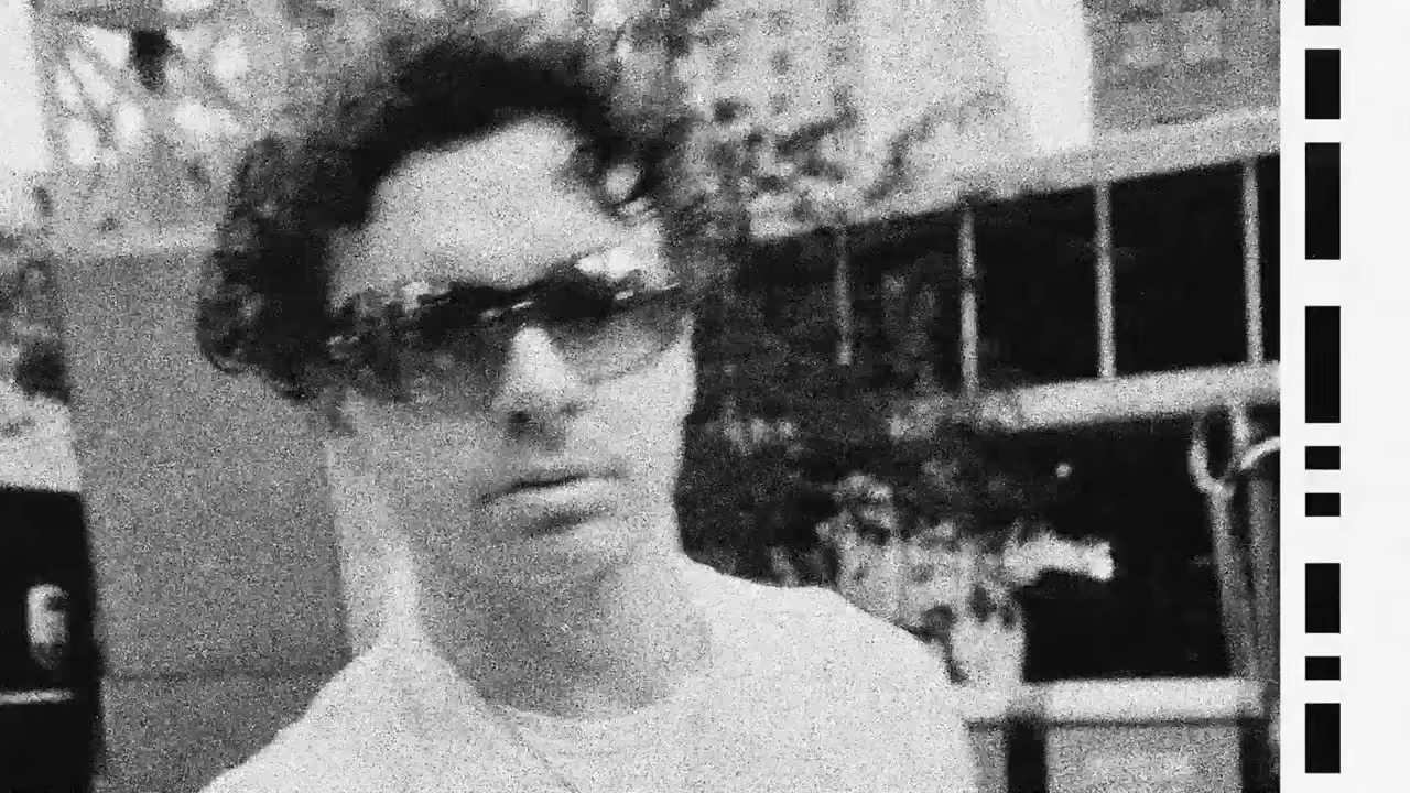 A person wearing sunglasses is captured in a grainy, black-and-white photo in an urban setting.