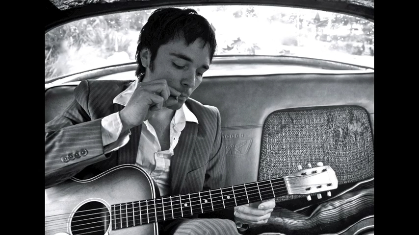 A musician sits in the backseat of a car playing an acoustic guitar, deeply engrossed in his music