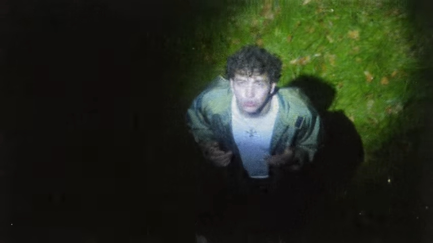 A young man stands outdoors at night, looking up at a light source. He is wearing a light jacket over a T-shirt