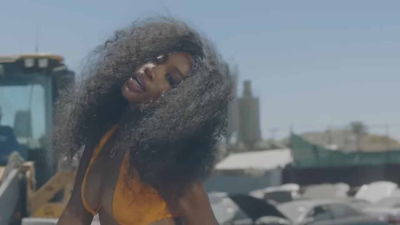 SZA poses outdoors in a revealing orange bikini top, with her hair voluminous and curly. Construction machinery and debris are visible in the background