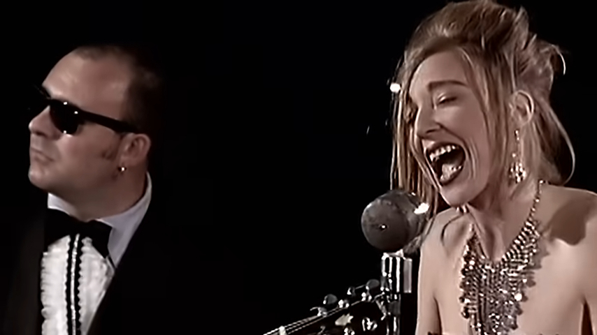 A man wearing sunglasses and a suit stands next to a woman with styled hair and a large statement necklace while she sings passionately into a microphone