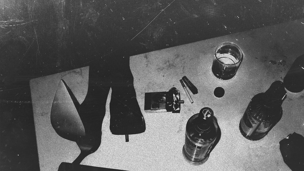 Close-up of a table with a high-heeled shoe, lighter, lipstick, candle, a glass of liquid, and an aerosol can scattered on it