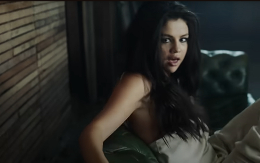 Selena Gomez sitting on a green couch, looking over her shoulder with a sultry expression, partially dressed in a loose tank top