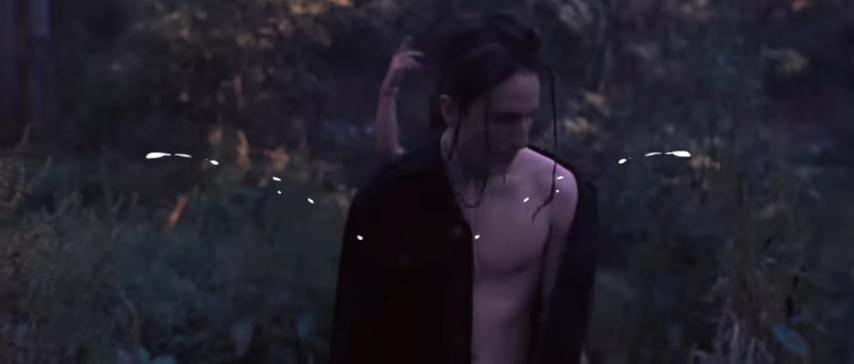Shirtless man in dark, nature setting, looking down with his hair tied back, wearing an open black jacket
