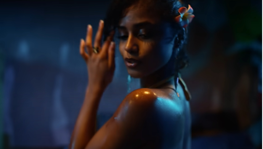 A woman wearing a flower in her hair poses seductively, illuminated by a spotlight in a sensual setting