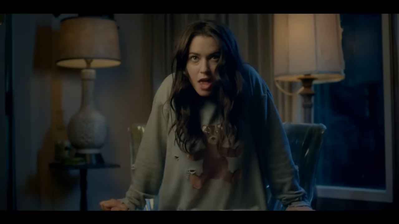 Woman in casual clothing looks surprised or shocked while leaning forward on a table in a dimly lit room with two lamps in the background