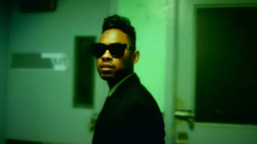 Miguel, wearing a dark suit and sunglasses, stands in a green-lit hallway