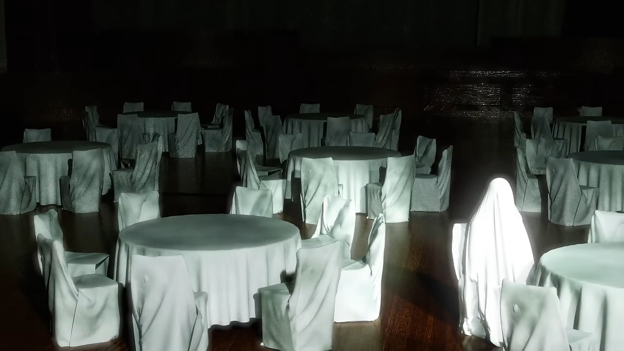 A dimly lit, empty room with round tables and chairs, all covered in white cloth. No persons are present in the image