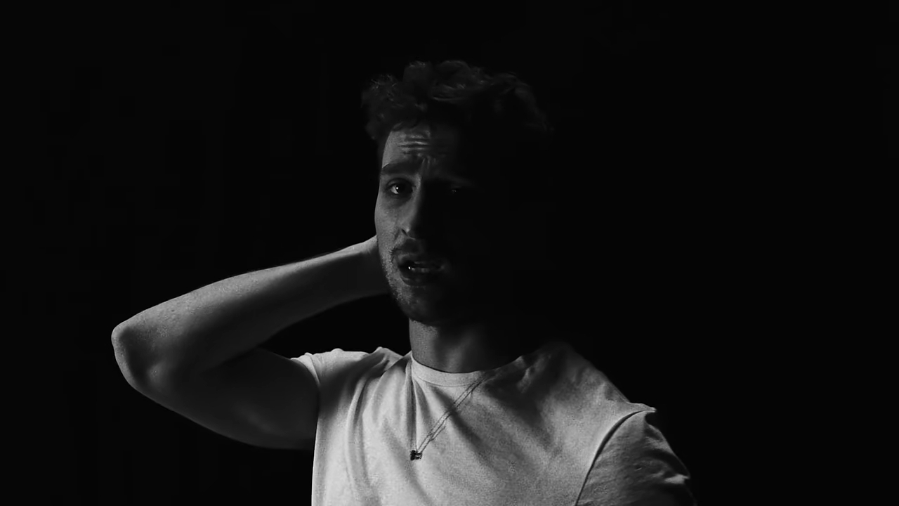 A man in a white T-shirt poses against a dark background, looking slightly distressed with one hand behind his neck