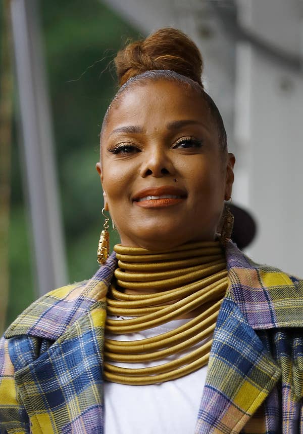 Janet Jackson, dressed in a plaid coat, top, and a statement necklace made of many layered rings, smiles for the camera