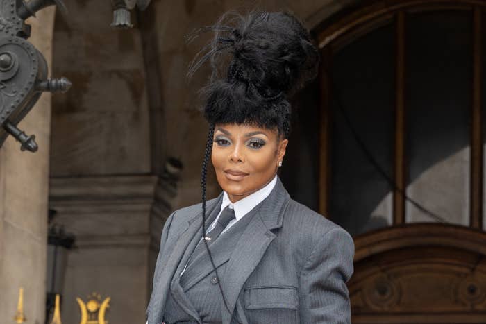 Janet Jackson in a stylish, tailored suit with an elaborate updo hairstyle, standing in front of an ornate building