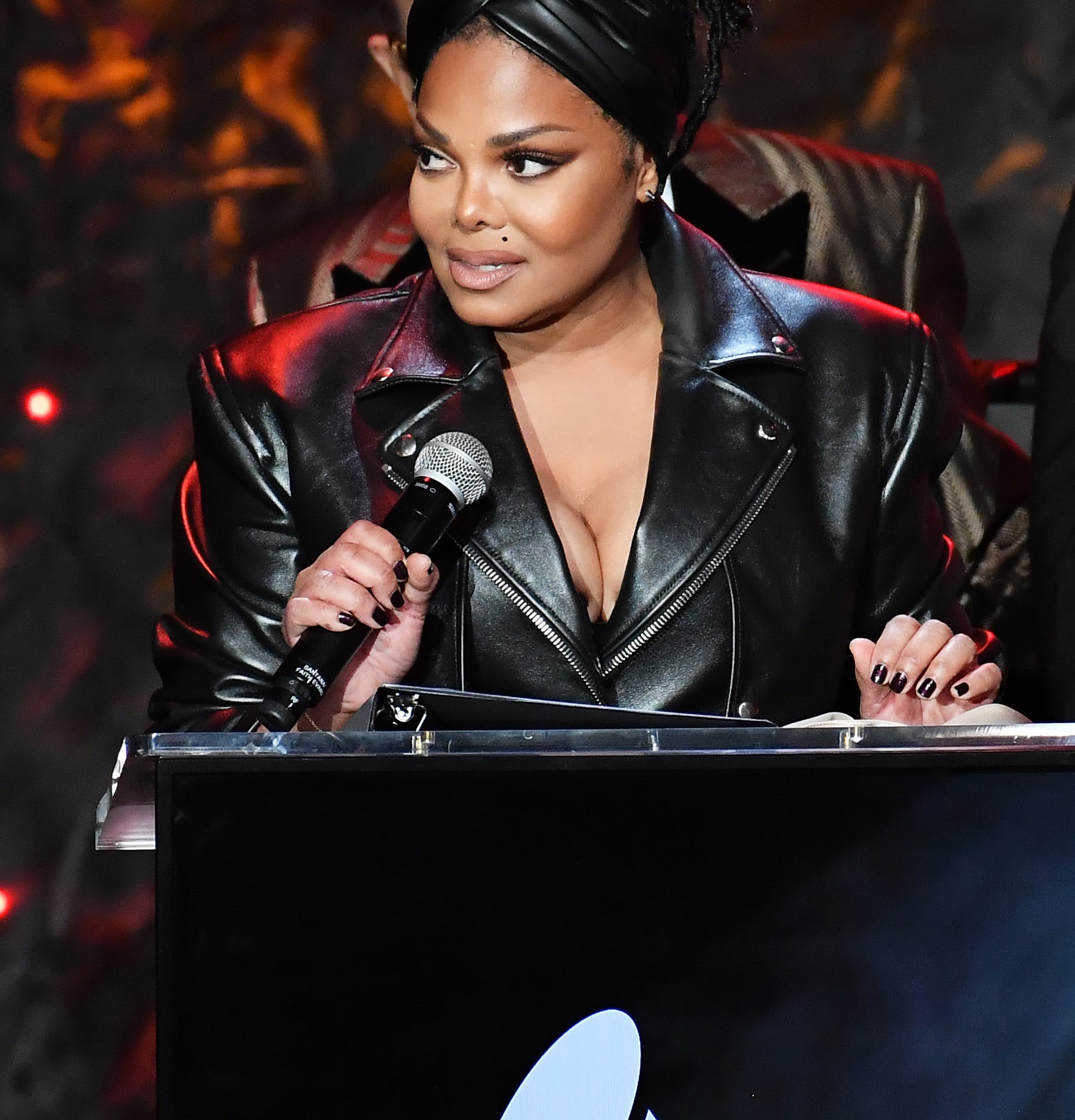 Janet Jackson stands at a podium with a Grammy logo, wearing a stylish leather jacket and a headwrap, speaking into a microphone
