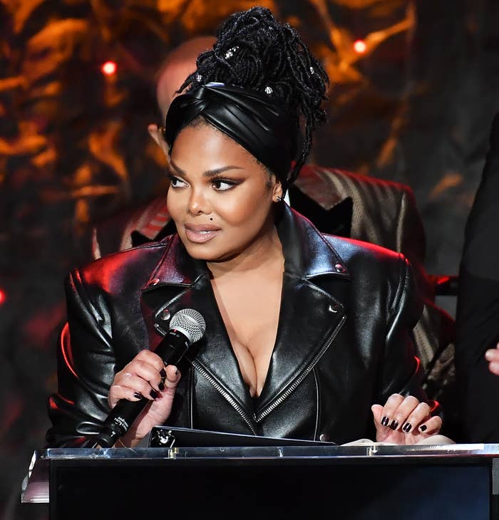 Janet Jackson stands at a podium with a Grammy logo, wearing a stylish leather jacket and a headwrap, speaking into a microphone