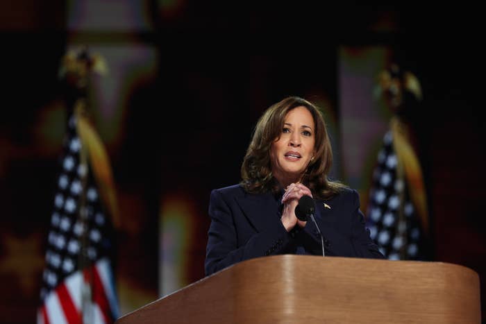 Kamala Harris stands behind a podium, addressing an audience with two American flags in the background