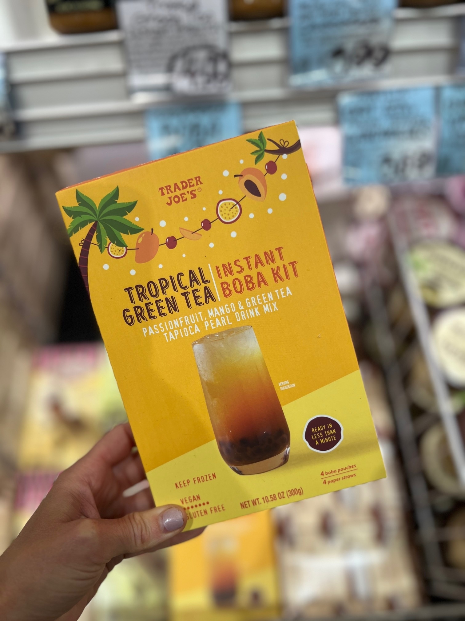 Hand holding a Trader Joe's Tropical Instant Green Tea Boba Kit box. Box text includes "Passionfruit, Mango, &amp;amp; Green Tea Tapioca Pearl Drink Mix."