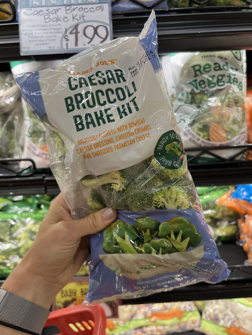A hand holding a bag of Trader Joe's Caesar Broccoli Bake Kit in a grocery store produce section. Sign shows price as $4.99