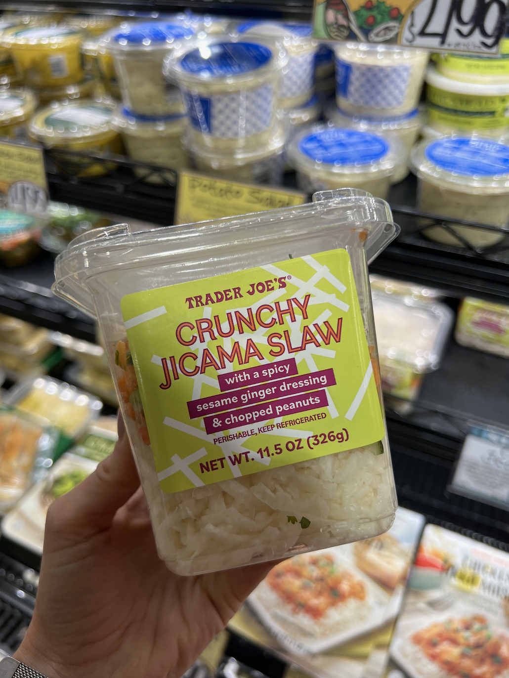 Hand holding a container of Trader Joe's Crunchy Jicama Slaw with a spicy sesame ginger dressing &amp;amp; chopped peanuts in a refrigerated grocery section