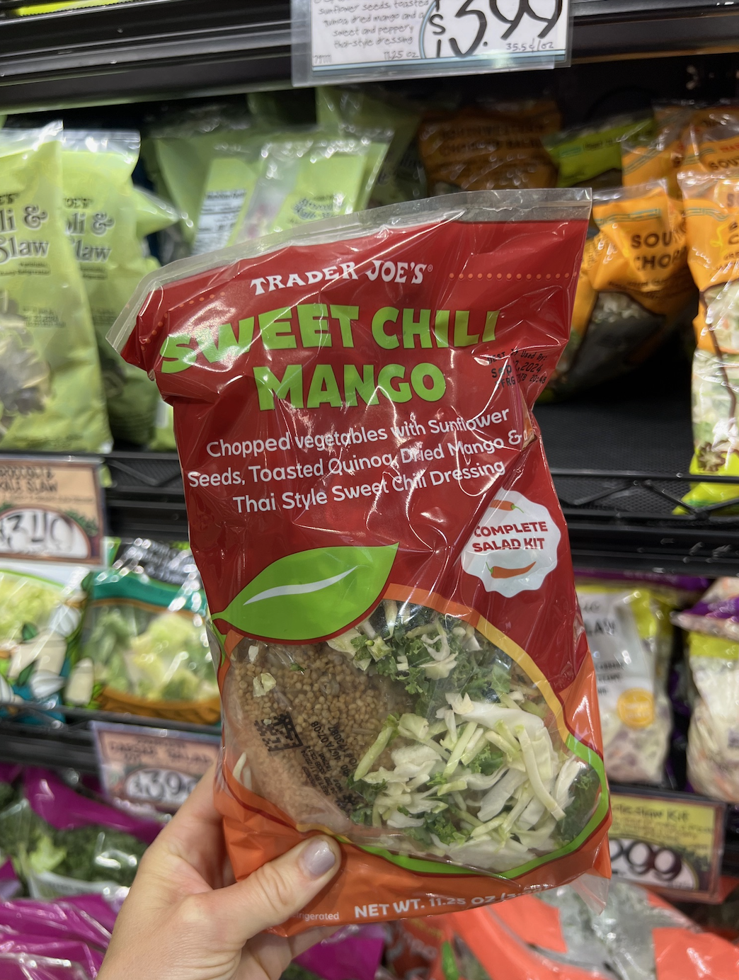 A hand holds a Trader Joe's Sweet Chili Mango salad kit in front of a supermarket shelf filled with various packaged salad items