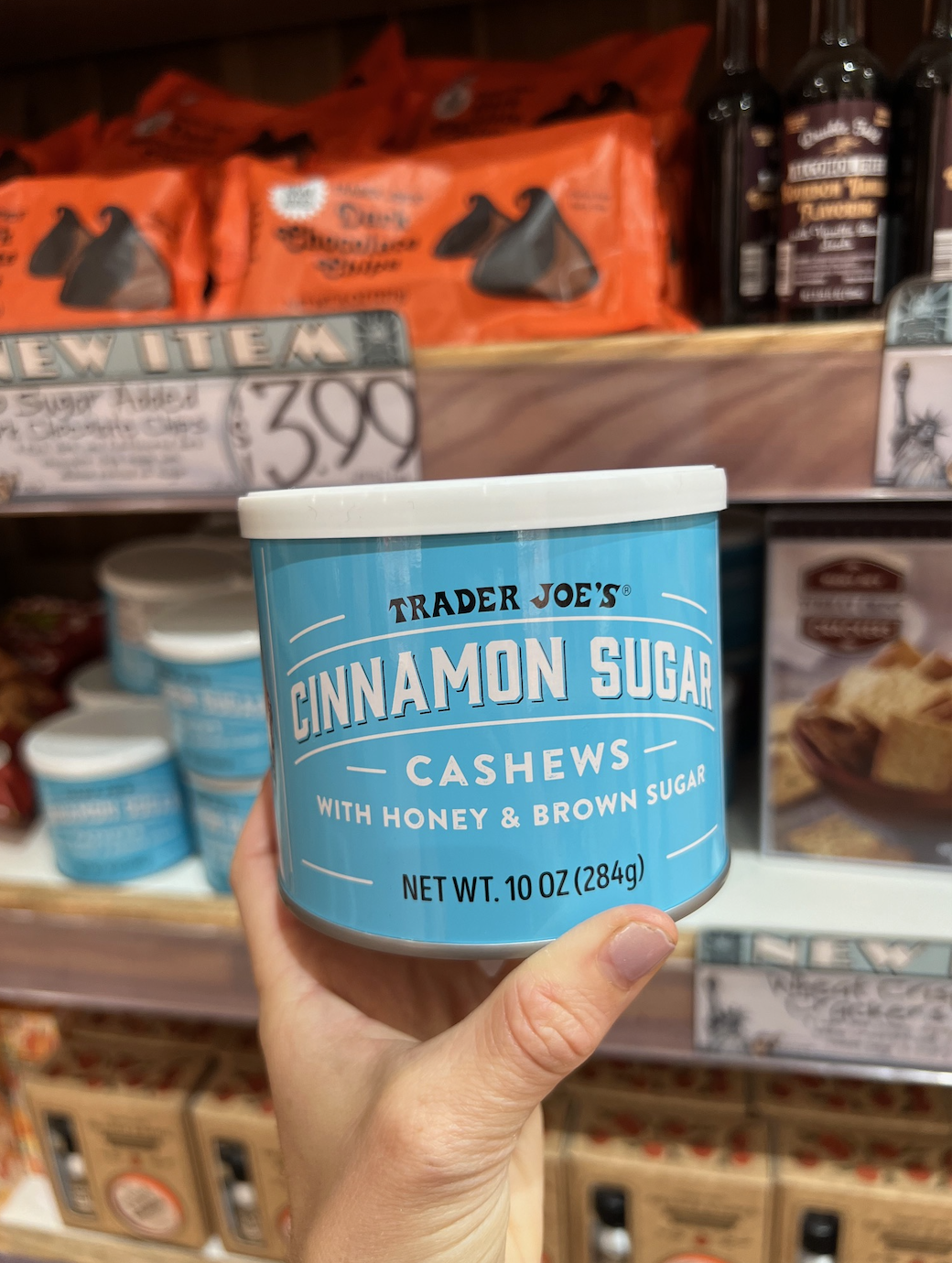 Hand holding a 10 oz container of Trader Joe's Cinnamon Sugar Cashews with honey and brown sugar in a grocery store aisle