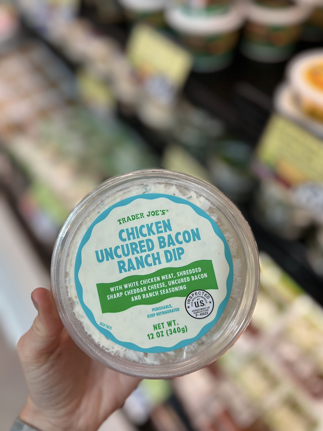 Hand holding a 12-ounce container of Trader Joe's Chicken Uncured Bacon Ranch Dip against a blurry store background