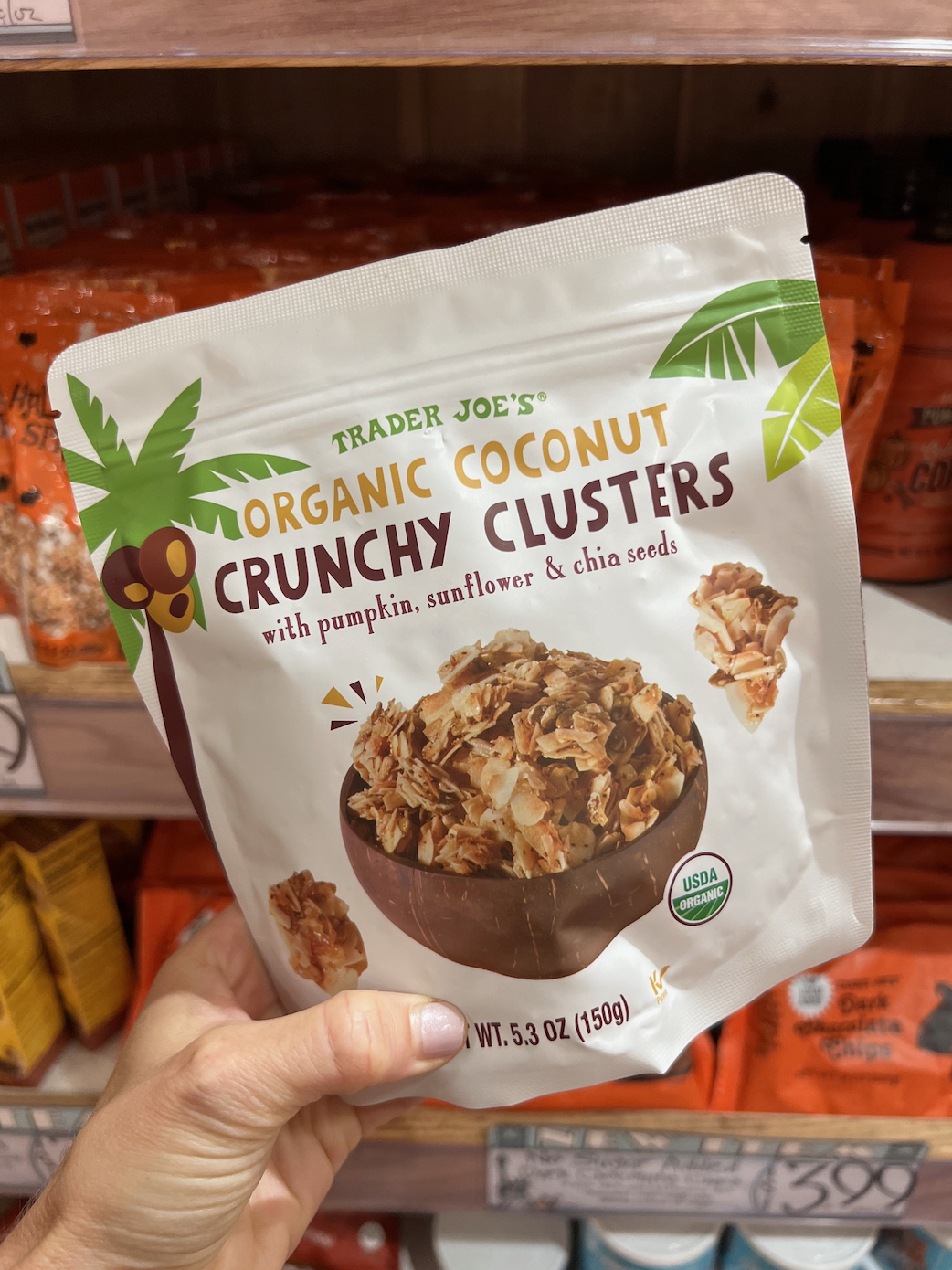 A hand holds a Trader Joe’s Organic Coconut Crunchy Clusters package with pumpkin, sunflower, and chia seeds, in a store aisle with similar products