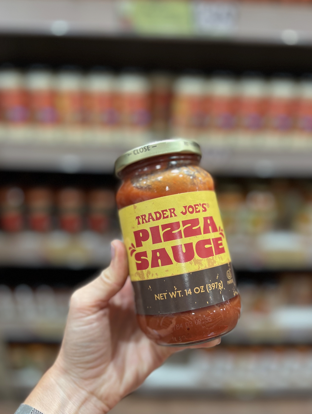 Hand holding a jar of Trader Joe's Pizza Sauce, labeled with a yellow and red design, in front of a store shelf filled with similar jars
