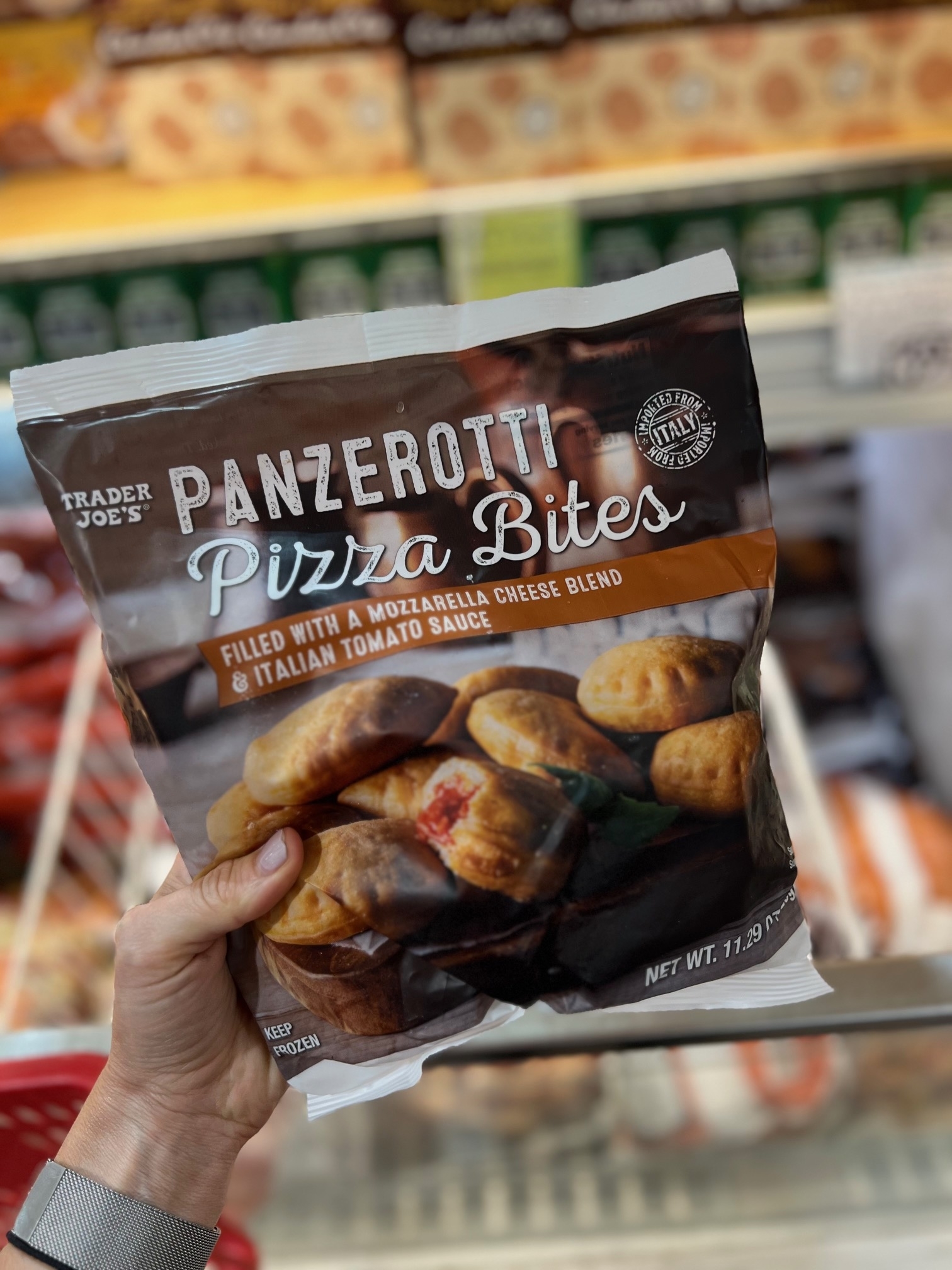 A hand holds a package of Trader Joe's Panzerotti Pizza Bites in a grocery store aisle. The packaging shows mini pizzas filled with mozzarella and tomato sauce