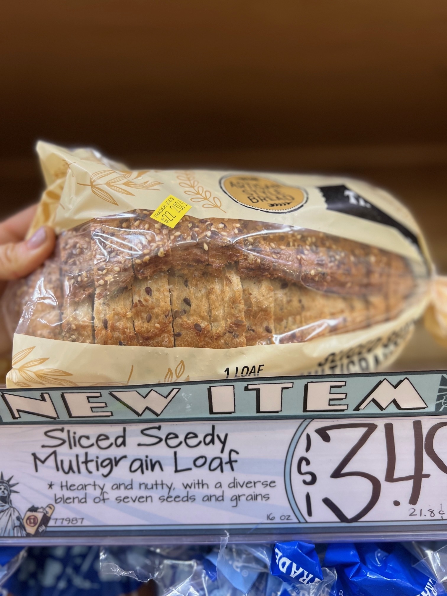 A hand holds a bag of sliced seedy multigrain loaf bread priced at $3.49, described as hearty and nutty with a diverse blend of seven seeds and grains