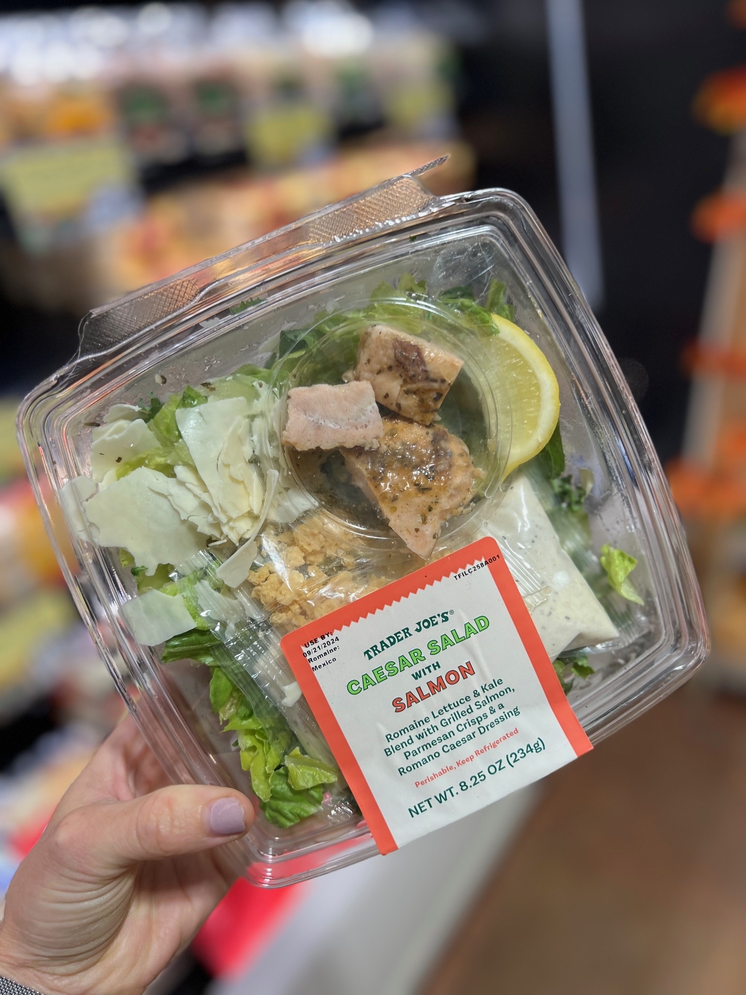 A hand holds a Trader Joe's Caesar Salad with Salmon in a plastic container. The label indicates it includes romaine lettuce, grilled salmon, and caesar dressing