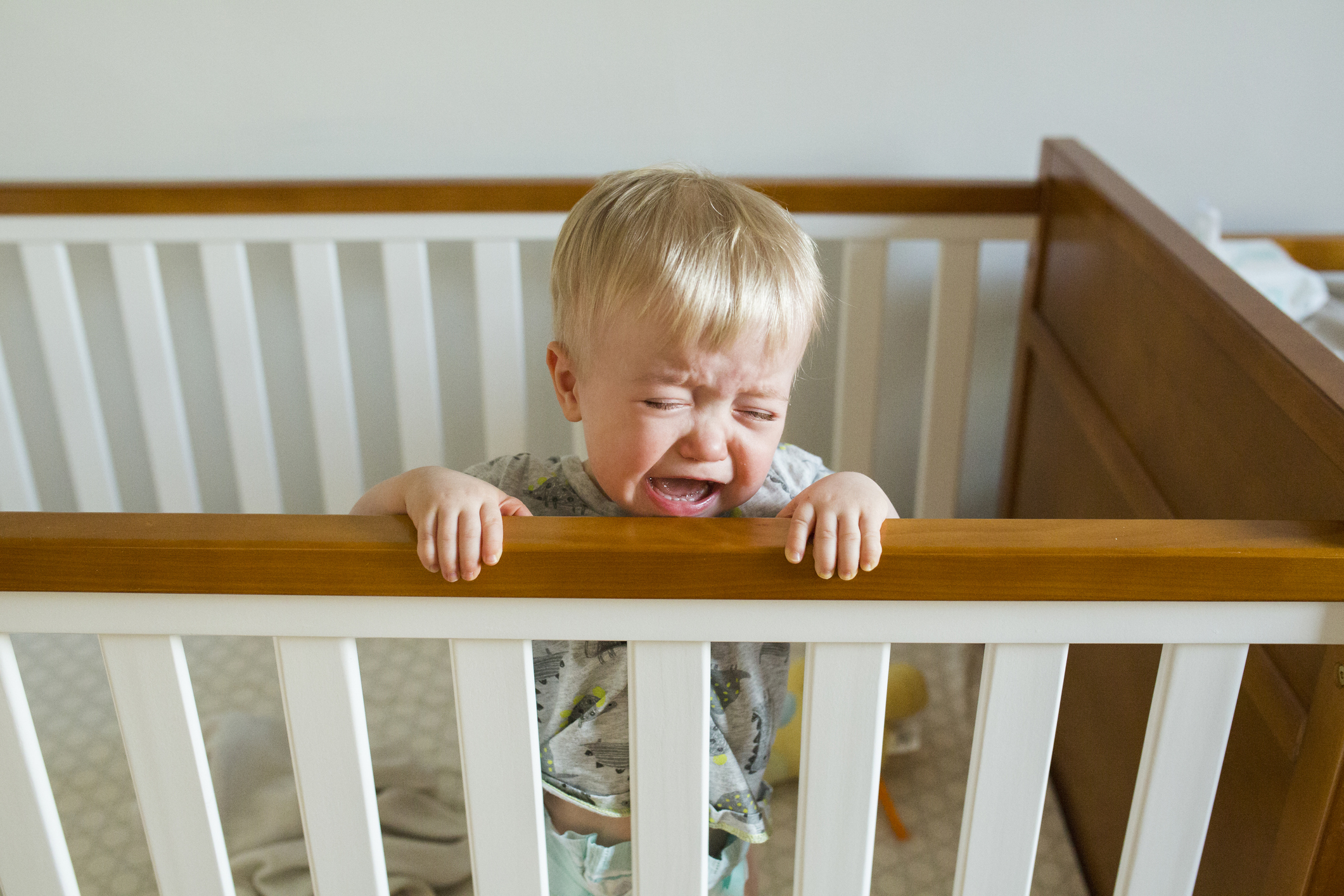 A crying blonde toddler standing in a crib, holding onto the edge with both hands
