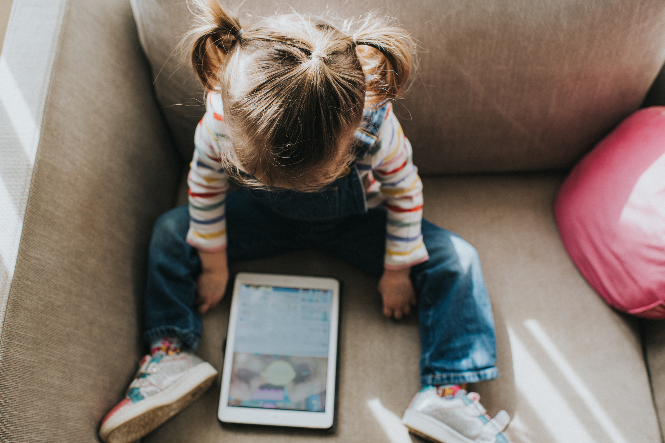 A young child with pigtails sits on a couch looking at a tablet. The child is wearing a striped shirt and jeans