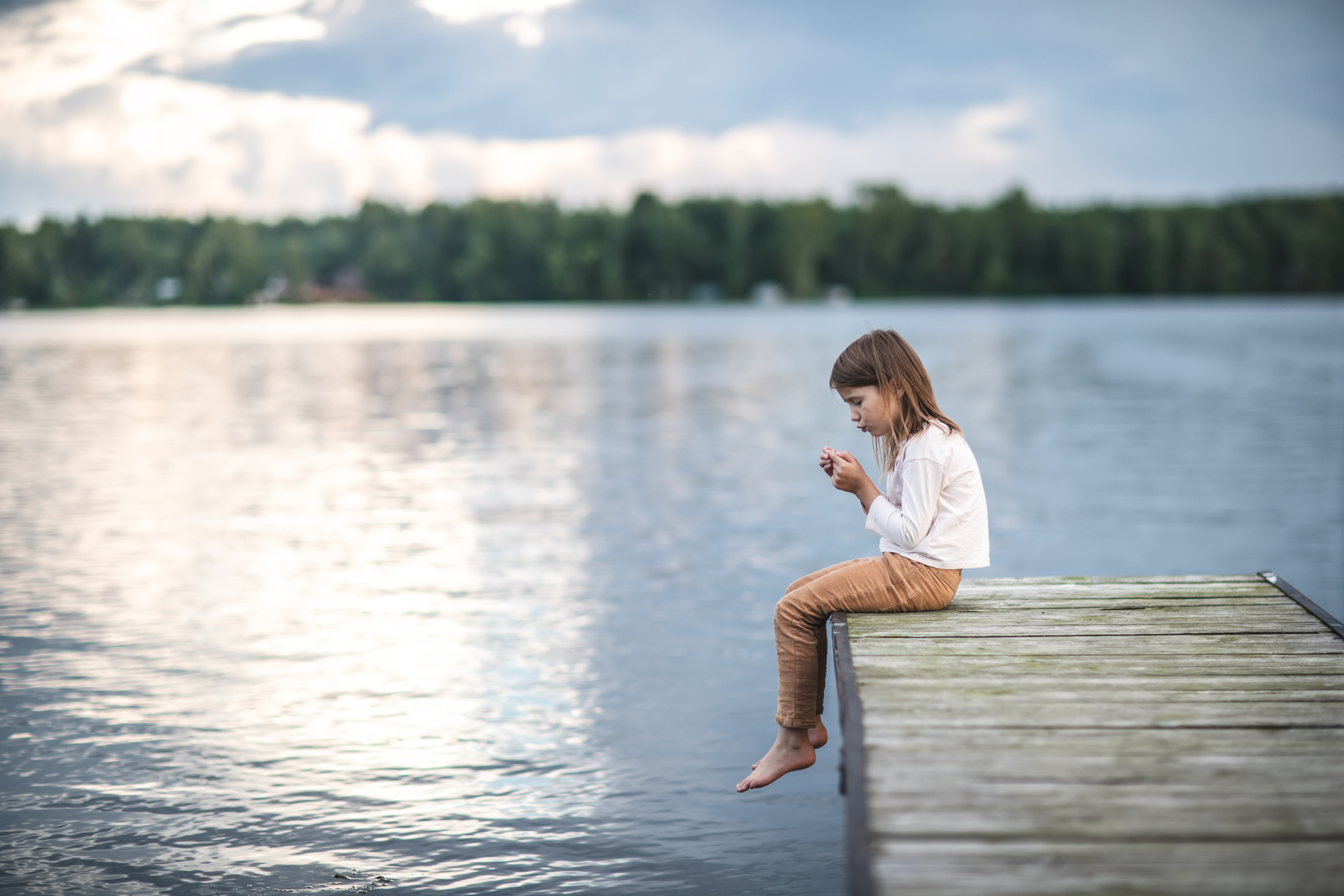 A child sits on a wooden dock, looking at something in their hands, with a lake and trees in the background