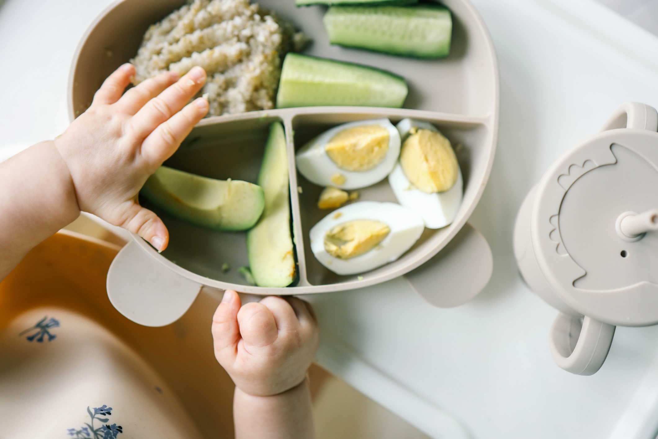 A baby's hands reach for food in a sectional plate containing avocado, hard-boiled eggs, cucumber sticks, and grains