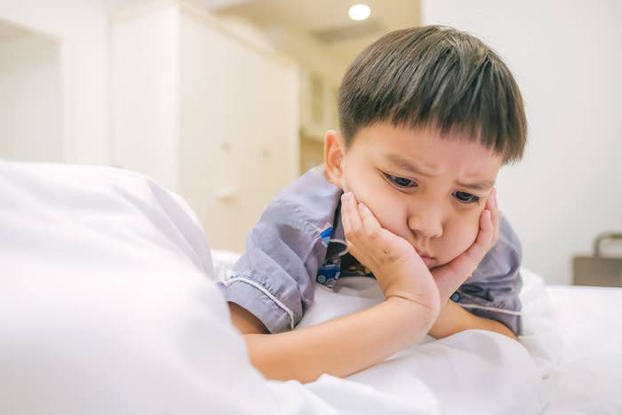 A young boy lies on his stomach on a bed, resting his head in his hands with a pensive expression. The background features a simple room