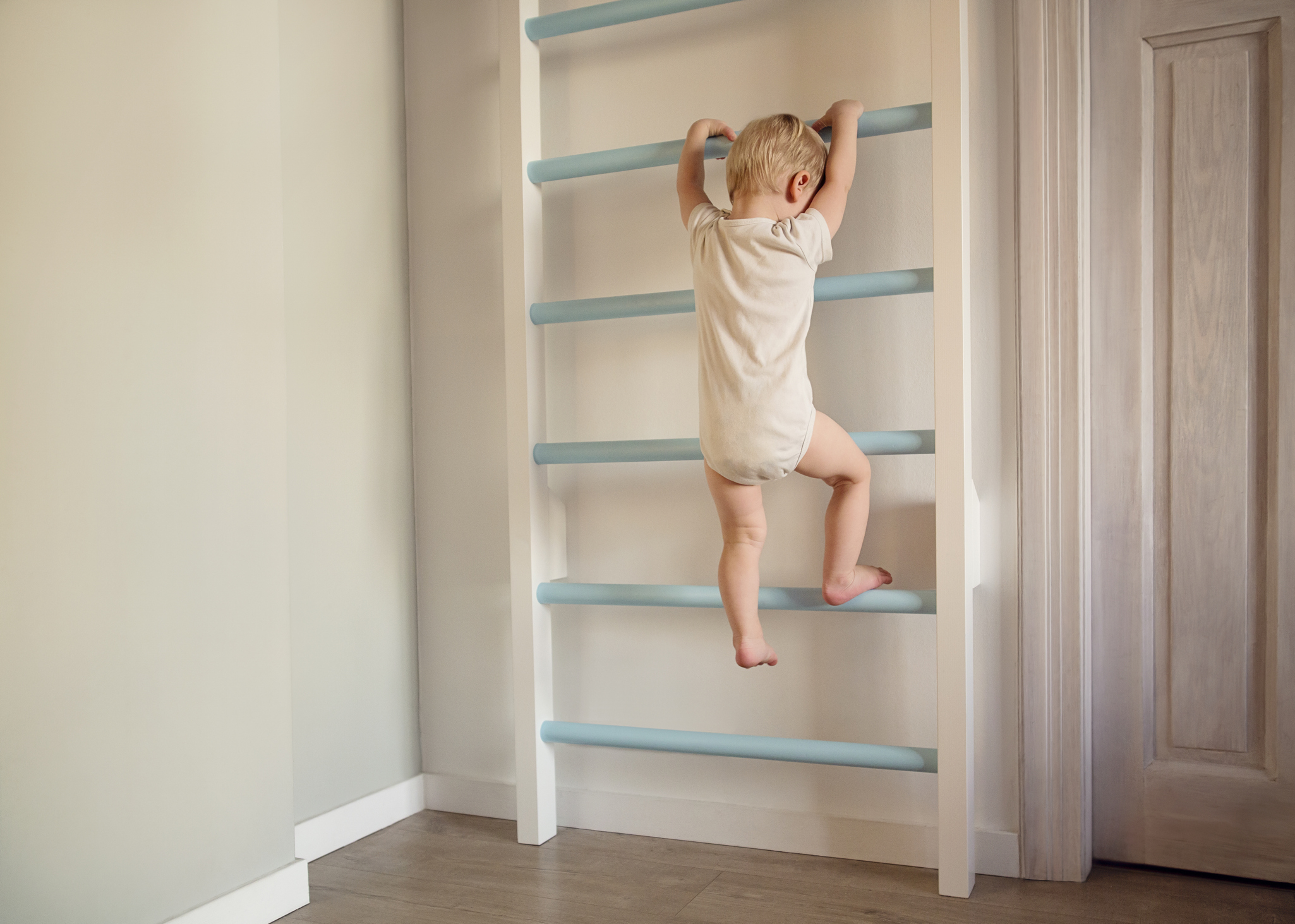 A young child climbs an indoor ladder attached to a wall, wearing a short-sleeved onesie. The room has a wooden floor and a door on the right