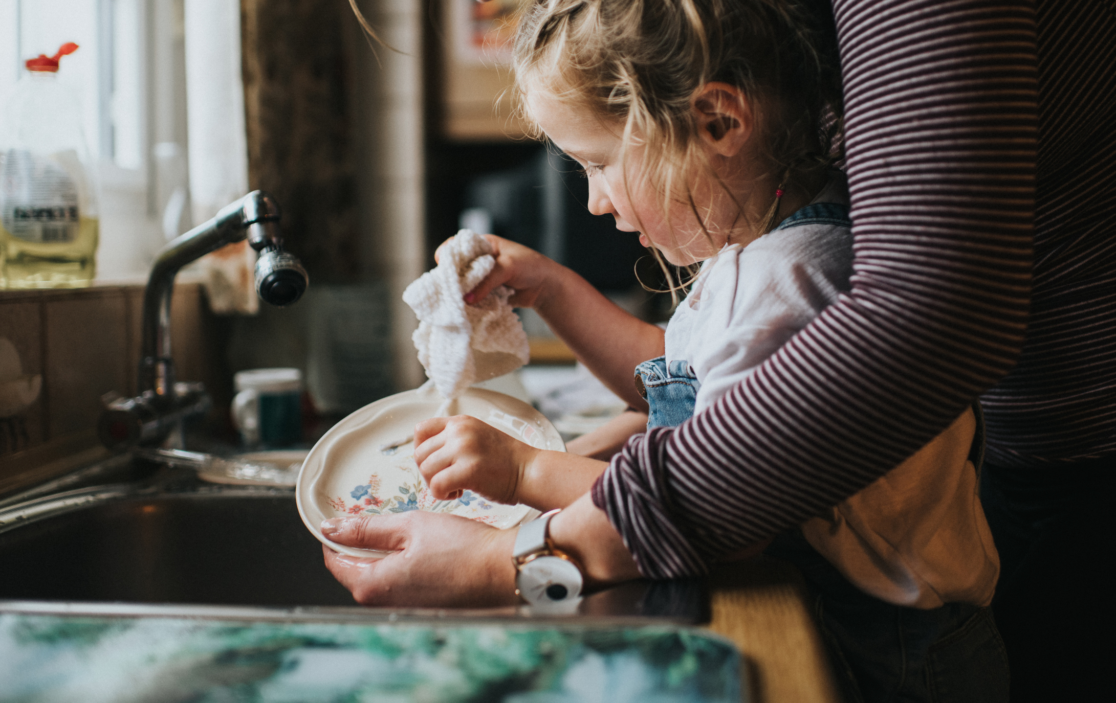 An adult helps a young child wash a plate at a kitchen sink. The child is focused on cleaning with a cloth while the adult gently guides them