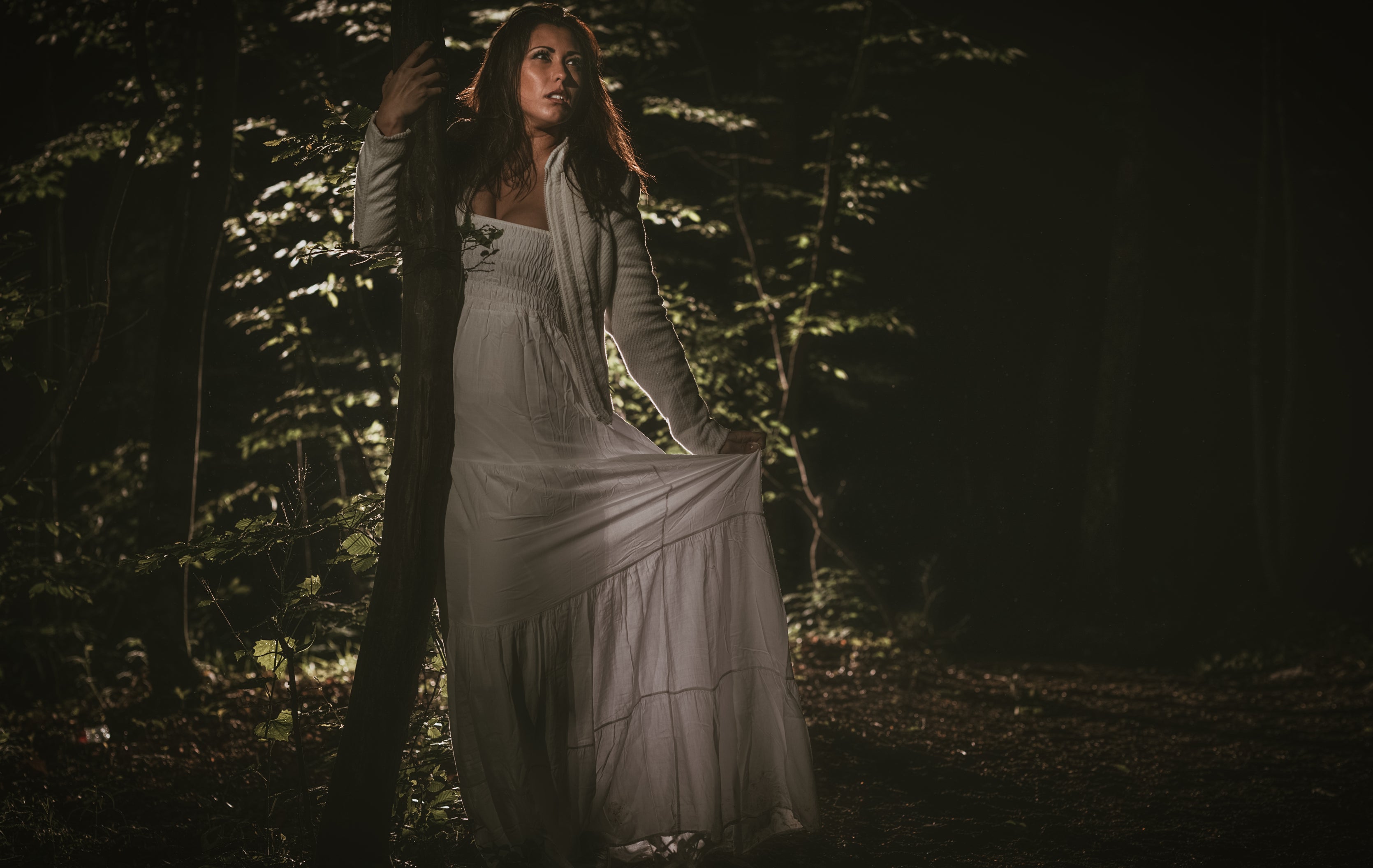 A woman in a flowing dress stands in a dark forest, illuminated by a light source, creating a mysterious and ethereal atmosphere