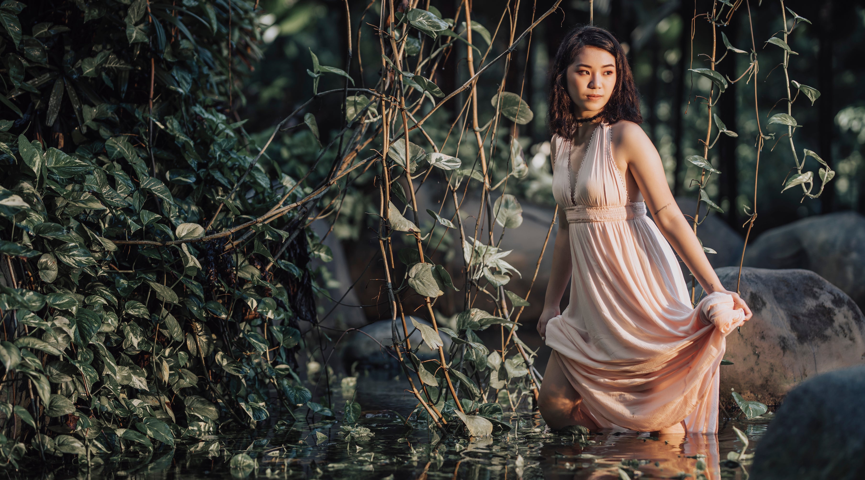 A woman in an elegant, flowing dress kneels in a lush, forest-like setting with water and greenery around her