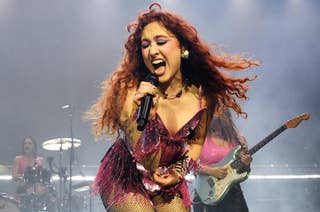 Tove Lo performing energetically on stage, wearing a fringed, sparkling outfit. A guitarist and drummer are playing in the background