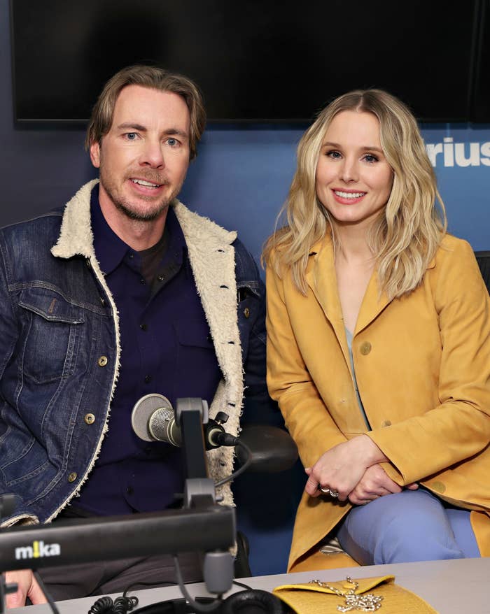 Dax Shepard and Kristen Bell sitting together in a radio studio, smiling at the camera. Dax wears a denim jacket and Kristen wears a coat