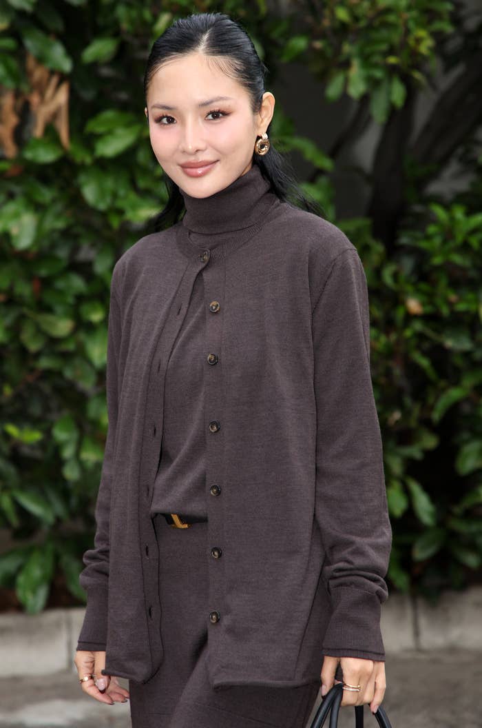 Eva Chen in a brown button-up cardigan over a matching short skirt, holding a black handbag, standing outdoors