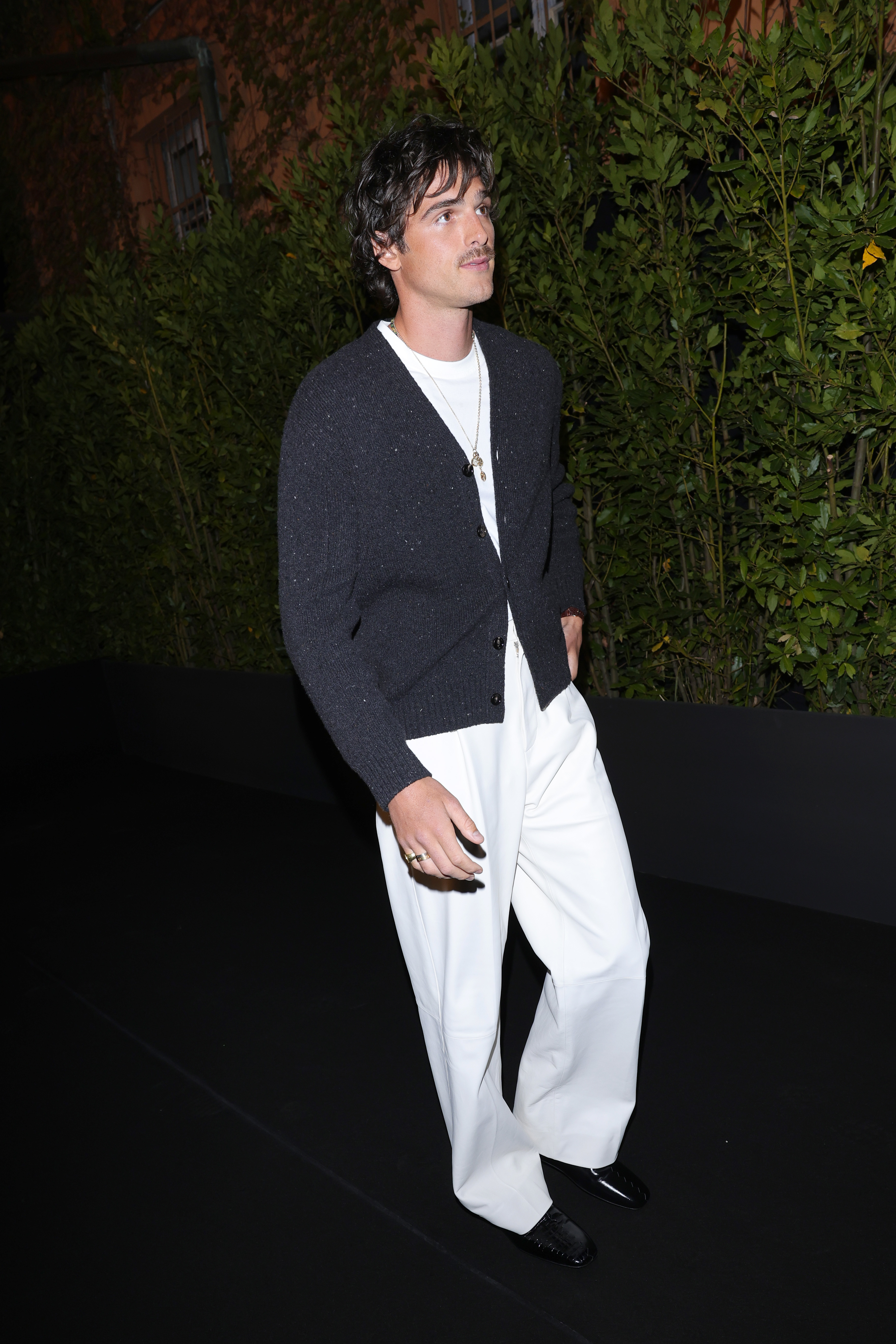 Jacob Elordi is wearing a black cardigan, white shirt, and white pants, walking outdoors in front of greenery