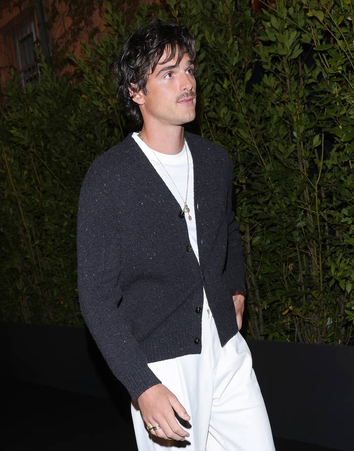 Jacob Elordi is wearing a black cardigan, white shirt, and white pants, walking outdoors in front of greenery