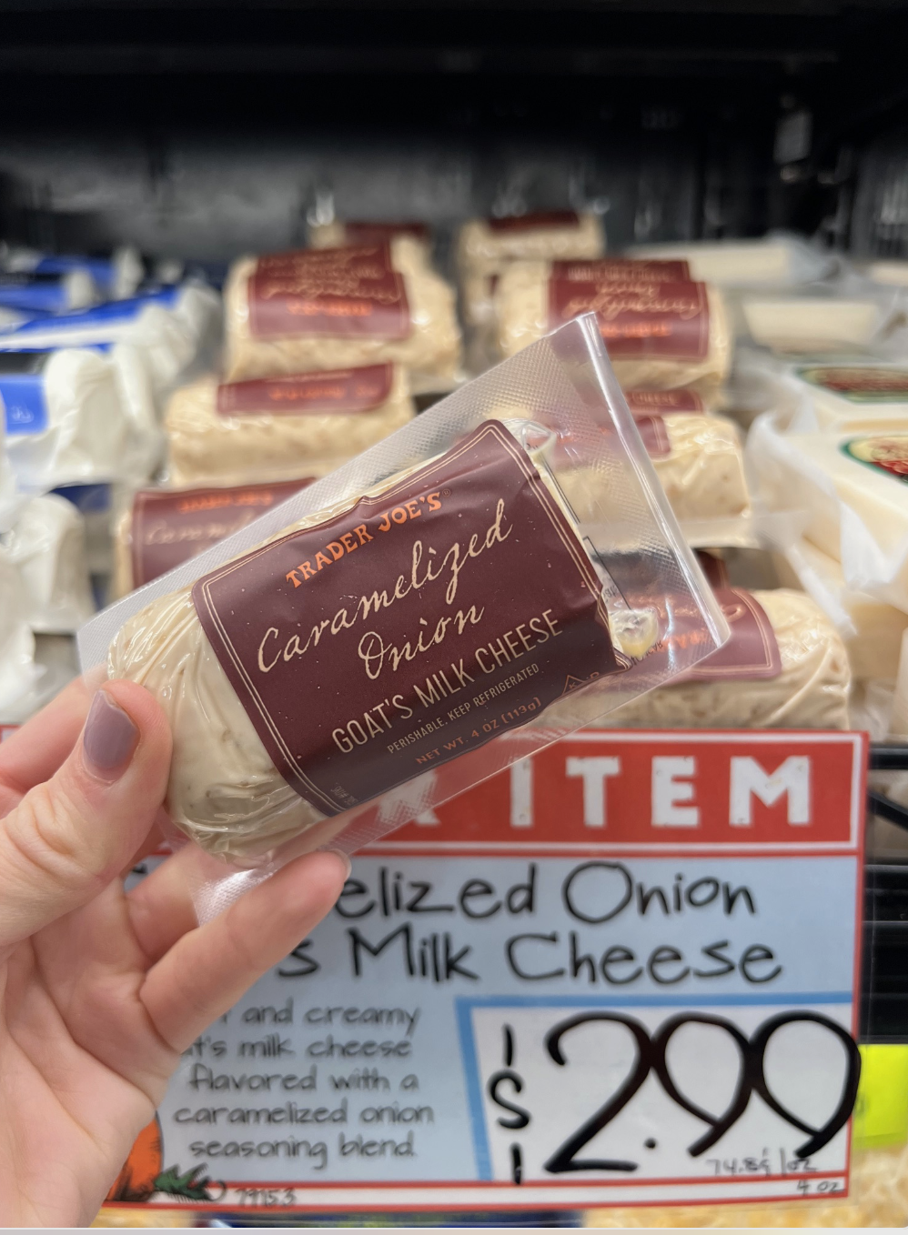 Hand holding Trader Joe's Caramelized Onion Goat's Milk Cheese in a store, with a price tag showing $2.99. Multiple cheese packs are displayed in the background