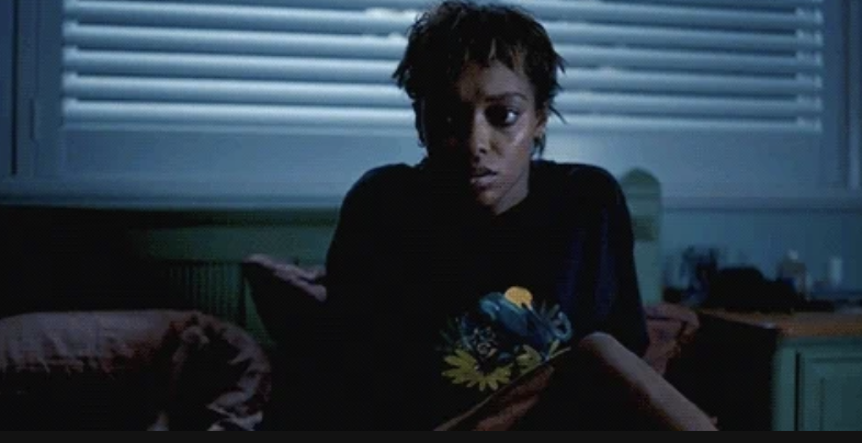 Person sitting on a bed in a dimly lit room, looking worried or thoughtful, wearing a dark shirt with a graphic design