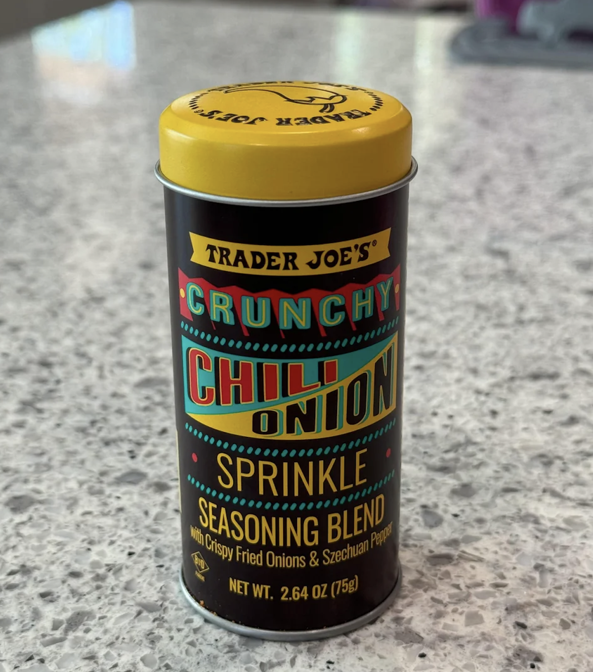 Trader Joe's Crunchy Chili Onion Sprinkle seasoning blend tin on a kitchen countertop. The tin promotes fried onions and Szechuan pepper, 2.64 oz